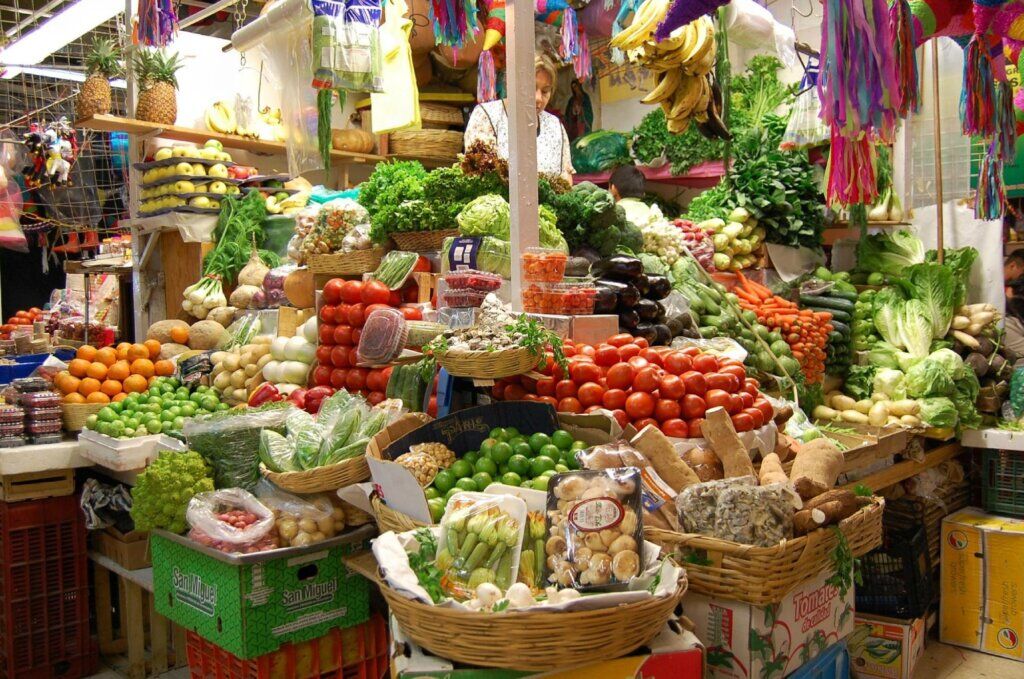 Produce stall piled with tomatoes, limes, greens, and fruit beneath hanging piñatas at the Coyoacán market