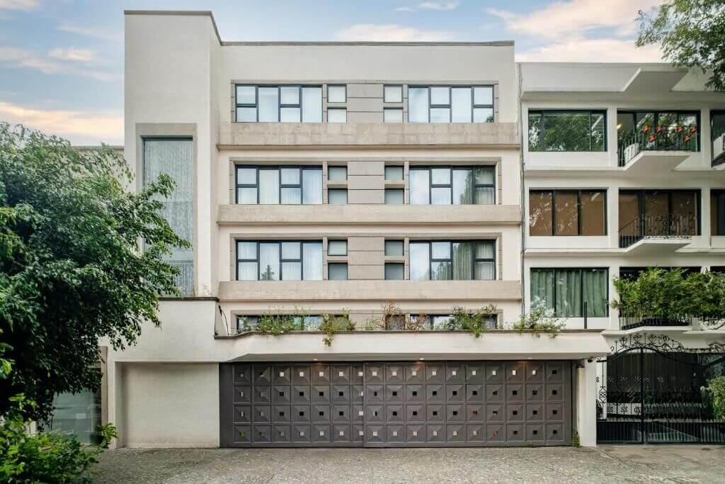 White mid-century apartment building with rows of windows, planted balconies, and a paneled garage door on a tree-lined street at District Polanco by Lumina