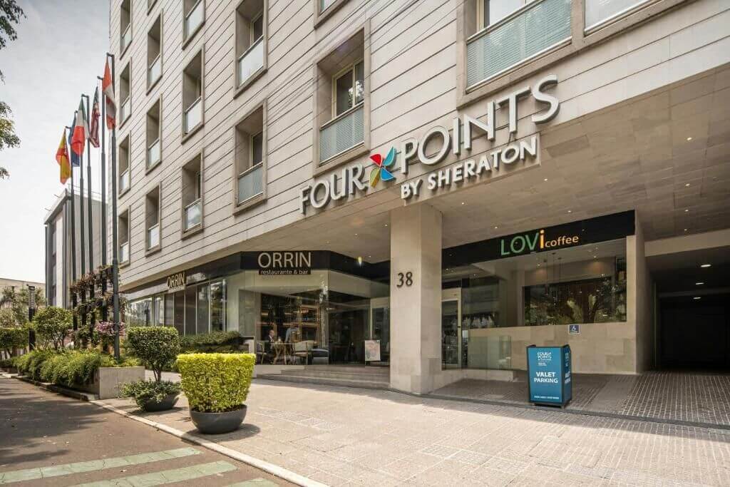 Street-level entrance of the Four Points by Sheraton with its signage, an on-site restaurant and coffee shop, potted plants, and flags in Mexico City