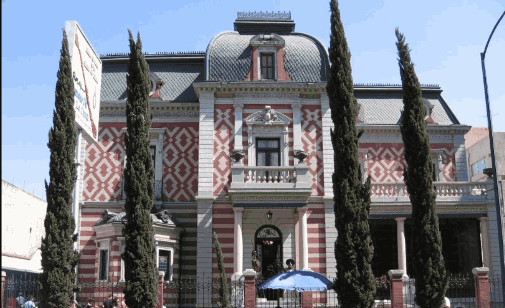 Red-and-white patterned French-style mansion housing the Museo de Cera behind tall cypress trees in Mexico City