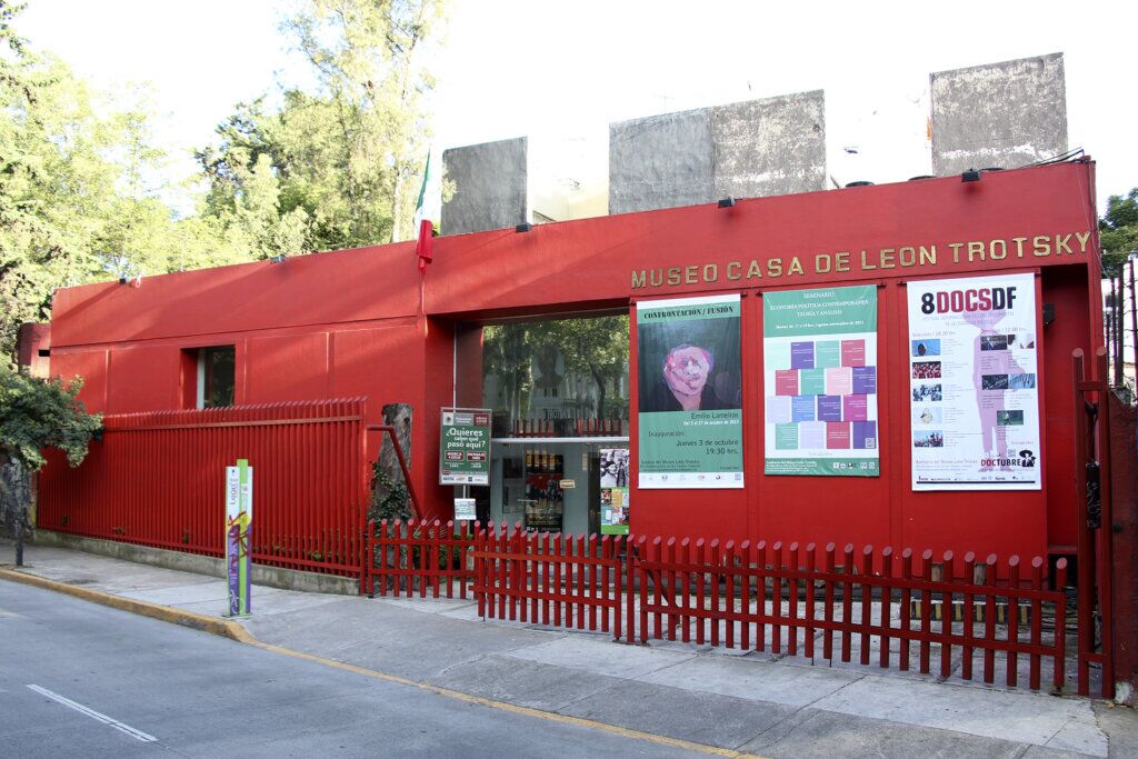 Red facade of the Museo Casa de León Trotsky with gold lettering, exhibition posters, and a red picket fence in Coyoacán