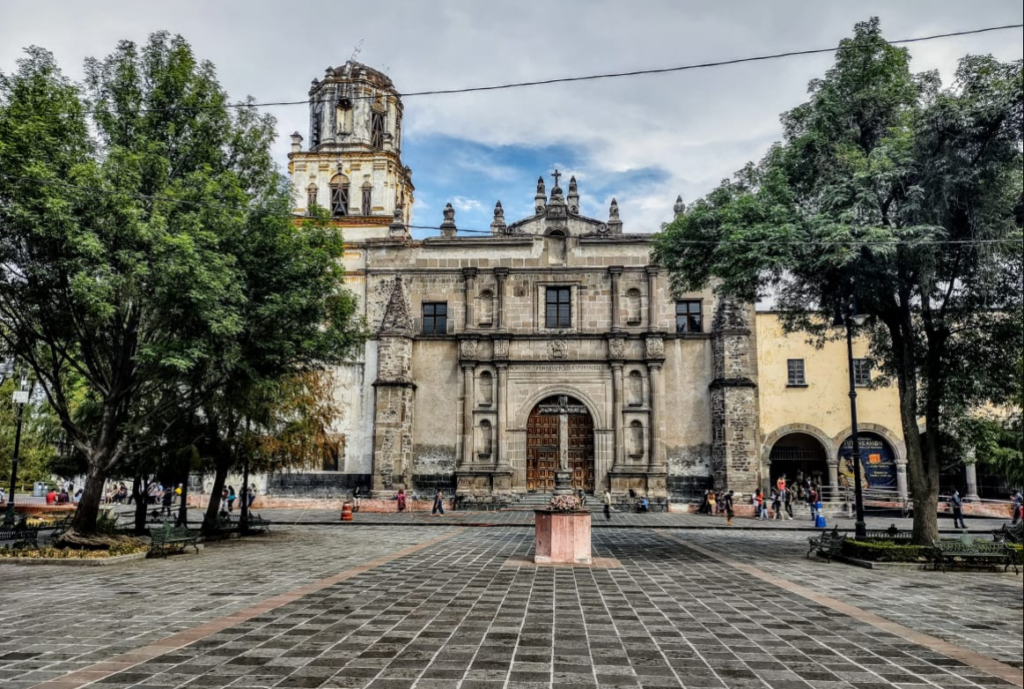 Stone baroque facade and bell tower of the Parroquia de San Juan Bautista seen across the plaza in Coyoacán