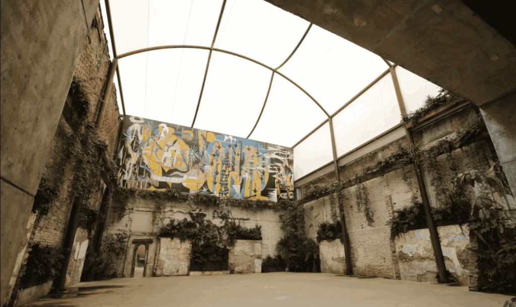 Restored industrial hall with exposed brick, a vaulted skylight, hanging greenery, and a large abstract mural at Proyecto Público Prim