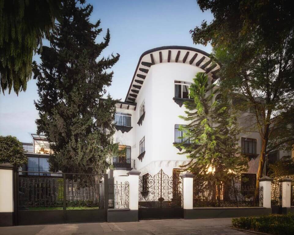 White colonial-style mansion with a curved roofline, dark trim, and wrought-iron gates behind tall pines at Pug Seal Allan Poe, Polanco