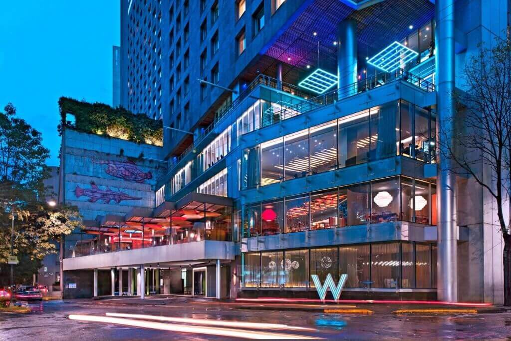 Illuminated glass facade of the W Mexico City at night, with a lit "W" sculpture out front and a rain-slicked street streaked with car lights